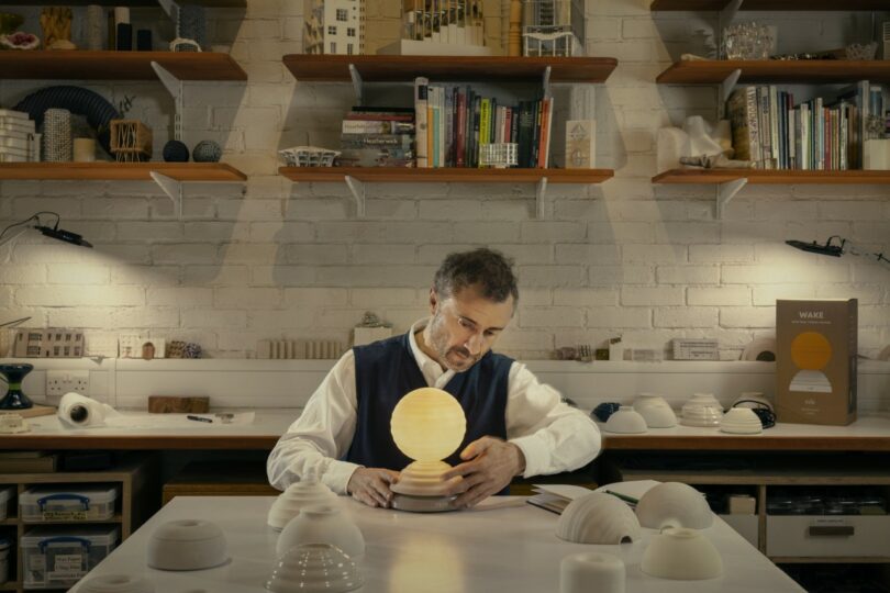 A man sits at a workbench in a studio, assembling a round lamp surrounded by ceramic pieces, with shelves of books and models in the background.