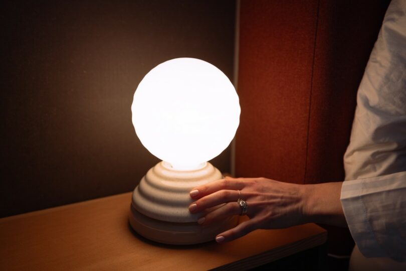 A person’s hand touches the base of a glowing round lamp on a wooden surface, with dark walls and a red panel in the background