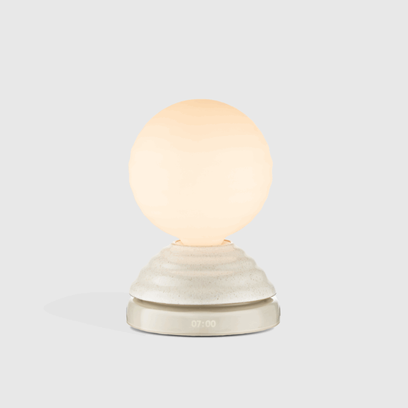 A spherical, softly glowing lamp sits on a textured, conical base with a digital time display reading "07:00" against a plain light gray background