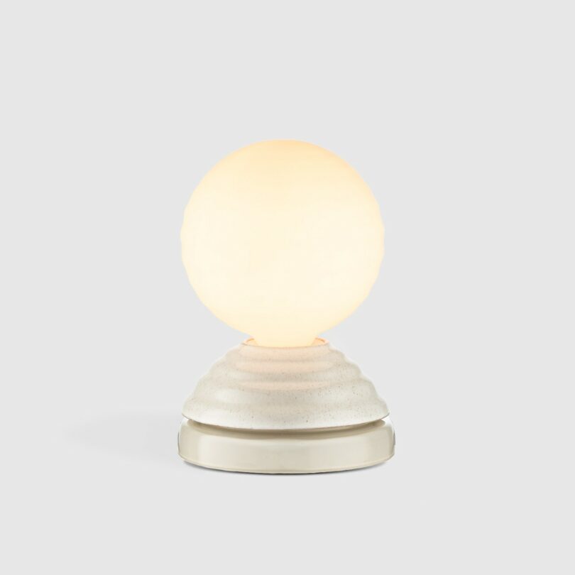 A small table lamp with a round frosted glass shade on a textured, light-colored ceramic base, photographed against a plain gray background