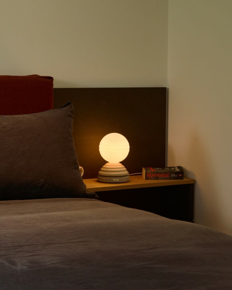 A neatly made bed with dark bedding, a bedside lamp turned on, a book, and a mug on a wooden nightstand against a plain wall
