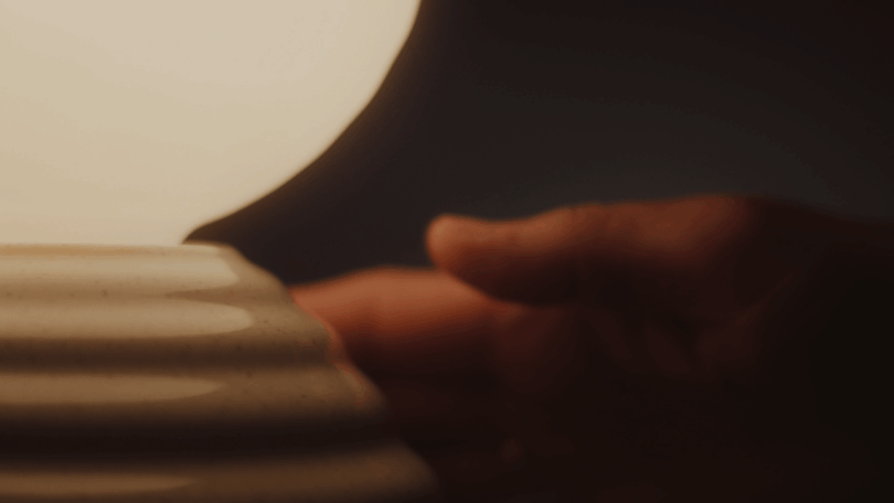 A hand reaches toward the base of a lit lamp, which has a ribbed ceramic surface, against a dark background