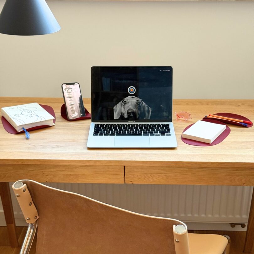 Una mesa de madera con un ordenador portátil, un perro, un teléfono, libretas, bolígrafos, notas adhesivas y una lámpara; se coloca una silla color canela frente a la mesa