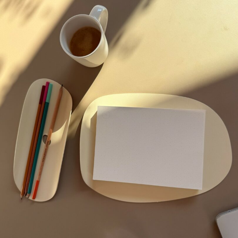Una taza de café, tres lápices de colores, un bolígrafo y un papel punteado en blanco sobre una bandeja de crema se encuentran sobre una superficie beige bajo luz natural