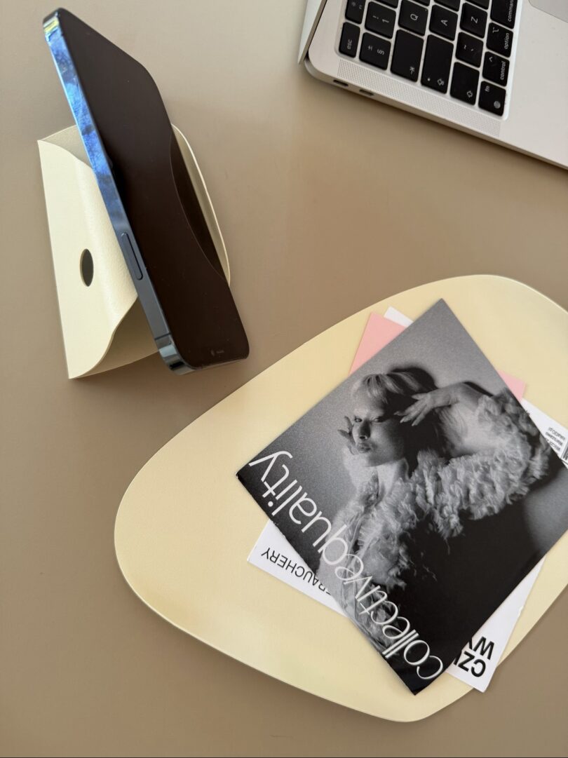 Sobre un soporte hay un teléfono inteligente, una computadora portátil, una bandeja con revistas y una fotografía en blanco y negro sobre una superficie beige