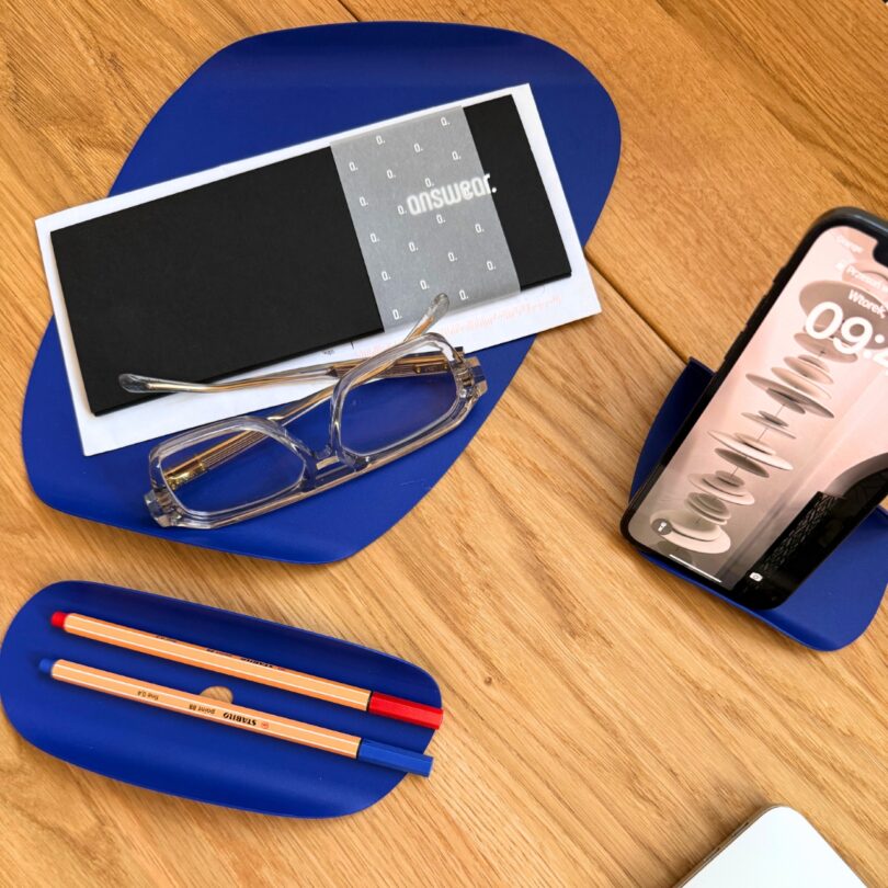 Gafas transparentes, bolígrafos, sobres cerrados, smartphone con pantalla visible y estuche de gafas azules sobre mesa de madera