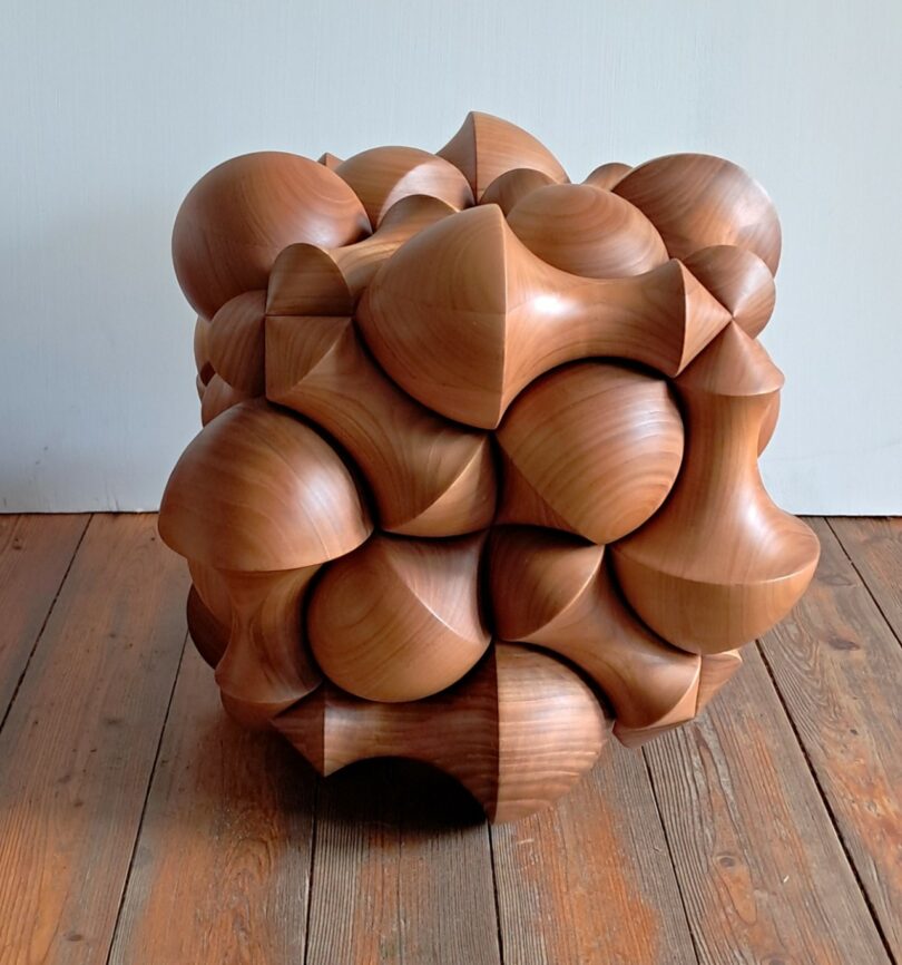 A wooden sculpture with an intricate geometric design featuring rounded, bulbous shapes, placed on a wooden floor against a plain white wall
