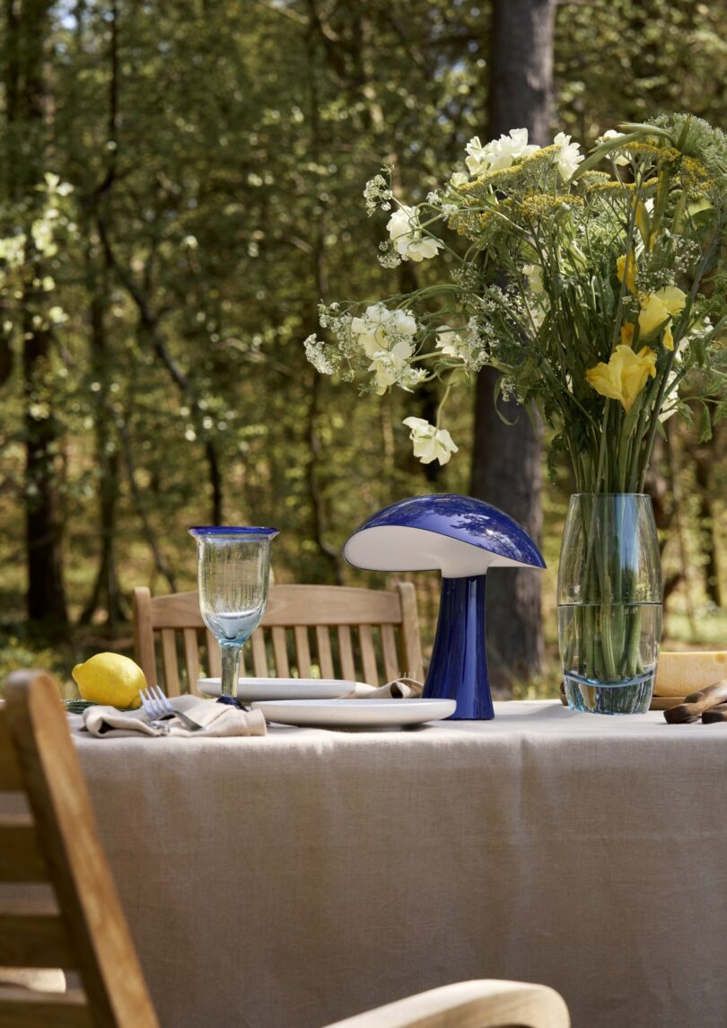 A wooden outdoor table set with plates, glasses, a vase of yellow and white flowers, a blue mushroom-shaped lamp, and a lemon, surrounded by chairs in a wooded setting
