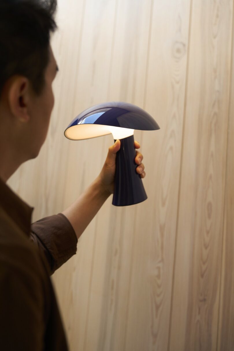 A person holds a modern, dark blue mushroom-shaped lamp that is turned on, in front of a light wooden wall
