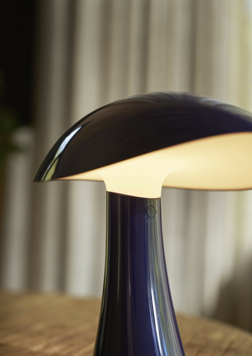 A close-up of a modern, dark-colored table lamp with a curved shade, turned on and placed on a wooden surface, with curtains blurred in the background