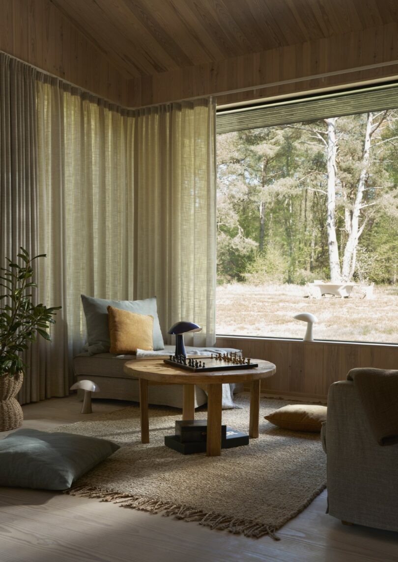 A cozy wooden living room with large windows, neutral-colored furniture, a chess set on a round table, and a view of trees outside