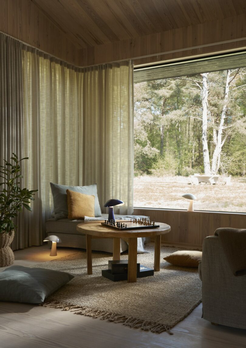 A cozy living room with neutral decor, a round wooden table set for chess, cushions on the floor, a large window, and a view of trees outside
