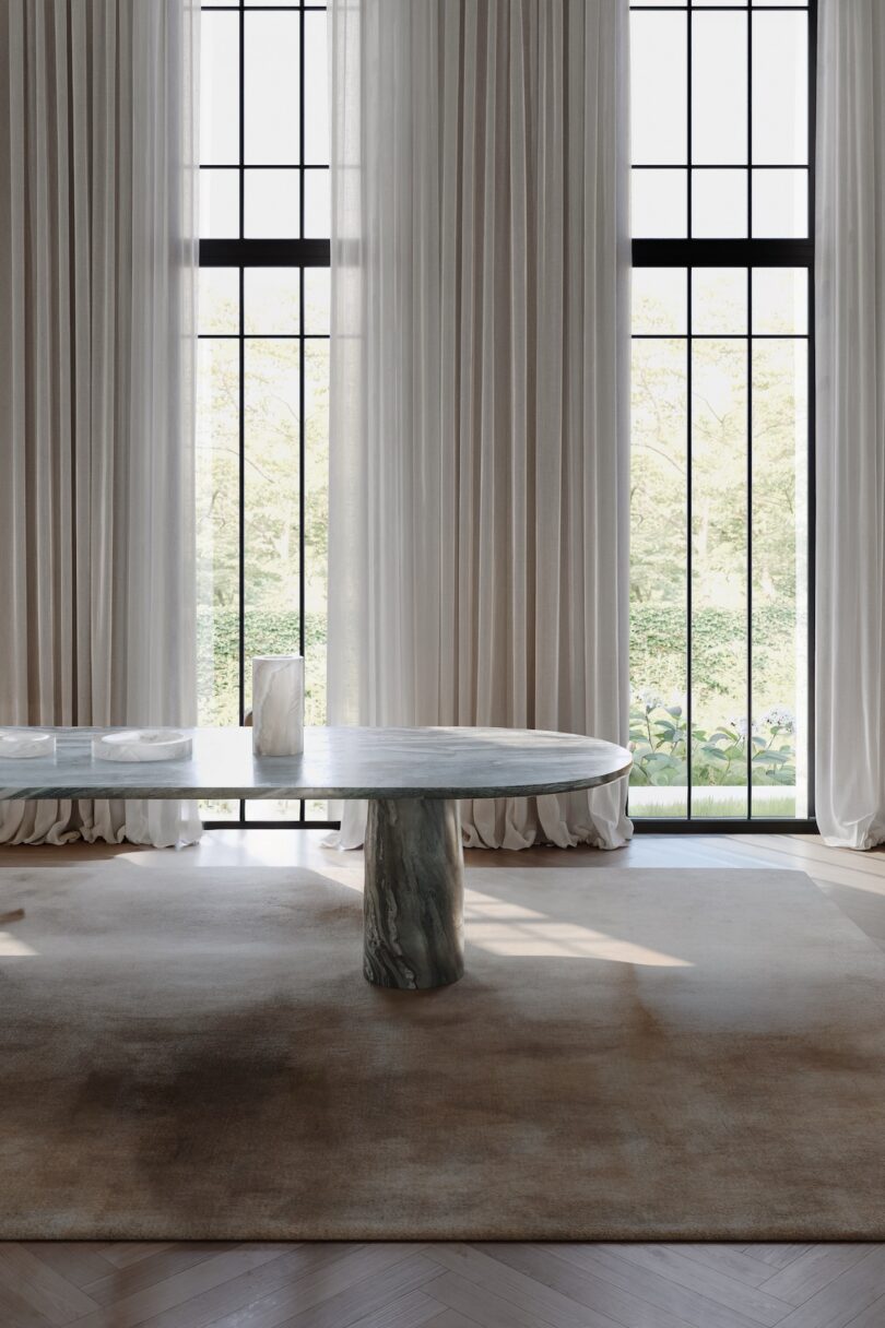 A marble table with a cylindrical base stands on a large rug in front of tall windows with sheer white curtains, letting in natural light.