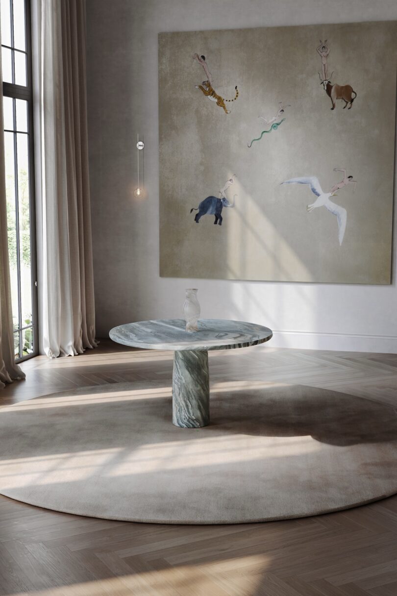 A round marble table with a glass vase sits on a circular rug in a minimalist room with tall windows and a large painting of animals on the wall.