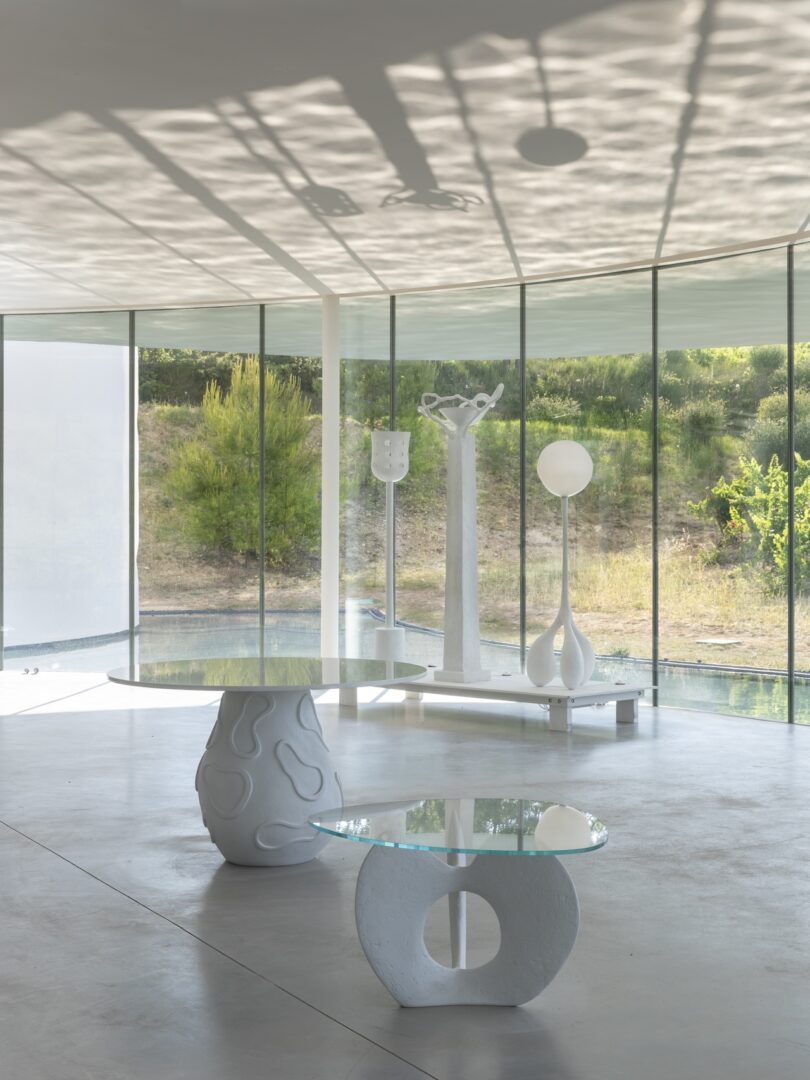 A modern interior with sculptural white furniture, two glass tables, and large floor-to-ceiling windows overlooking greenery.