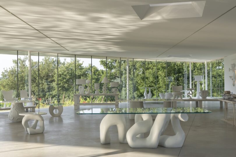 Minimalist room with large glass walls, featuring sculptural white furniture and decor, and views of green trees outside.