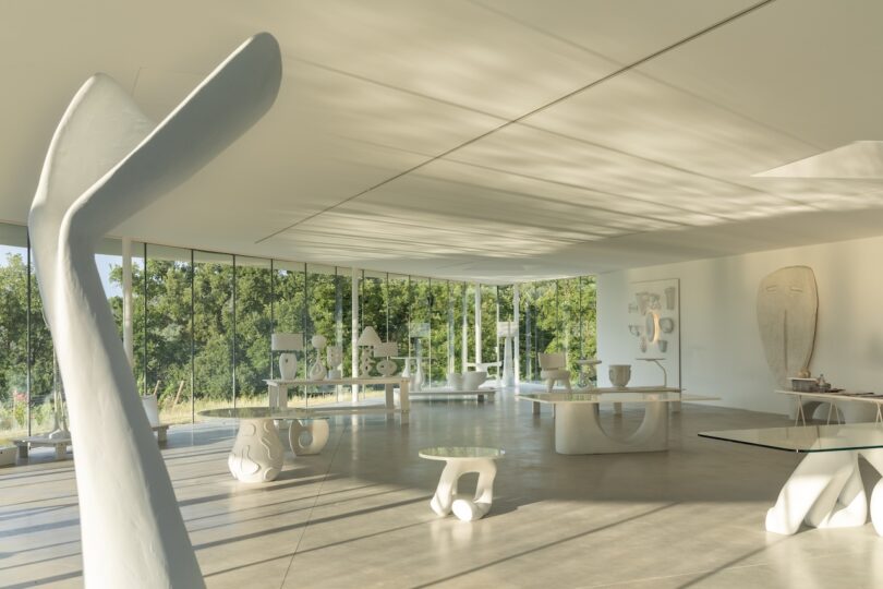 A modern, sunlit gallery with large windows displays abstract white sculptures and furniture on a polished concrete floor. Trees are visible outside.