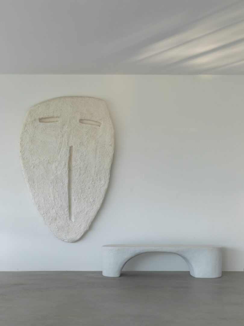 Large, abstract white face sculpture mounted on a plain wall above a minimalist, white, curved bench in a modern, empty room.