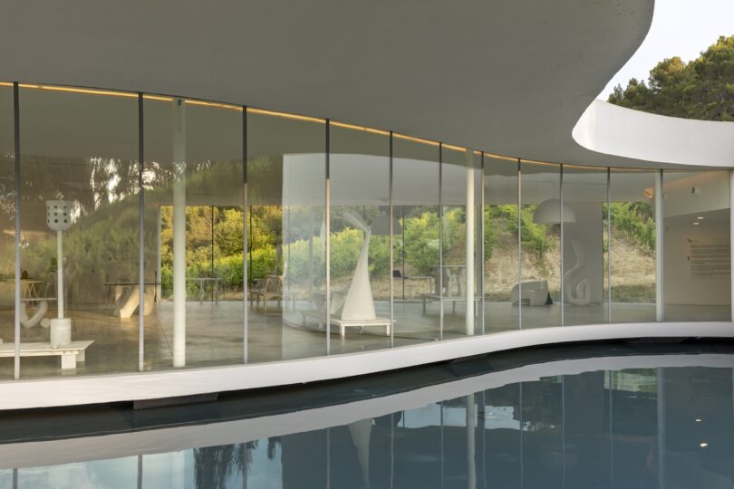 A modern building with floor-to-ceiling glass walls overlooks a curved pool, with minimalist white furnishings and greenery visible through the windows.