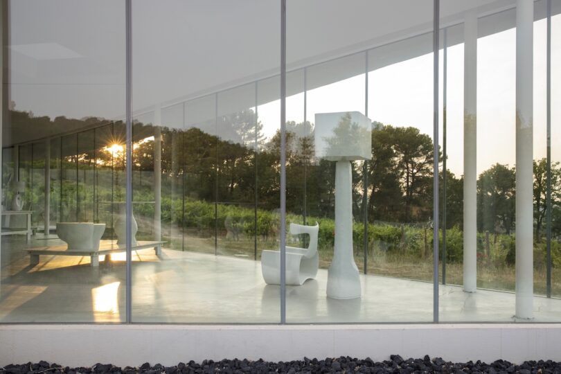Modern glass-walled room with white minimalist furniture, overlooking trees and greenery outside during sunset.