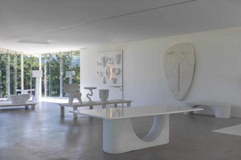 Minimalist room with white sculptural furniture, large abstract face wall art, and floor-to-ceiling windows overlooking greenery.