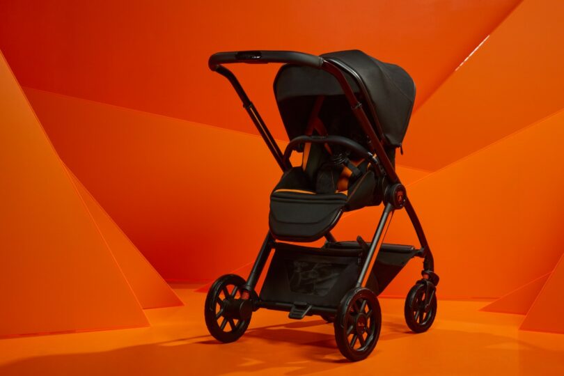 A modern black Silver Cross baby stroller is displayed against an orange geometric background, evoking the sleek sophistication of a Lamborghini