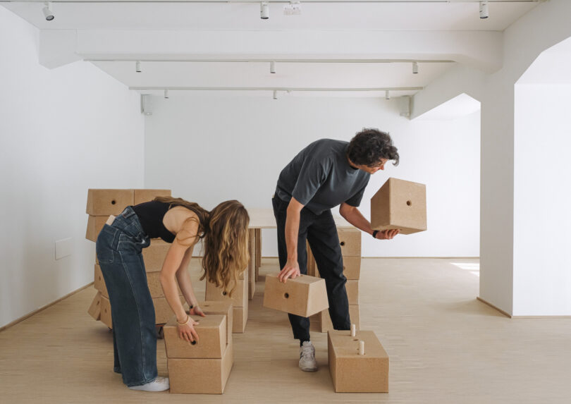 Two people are assembling or stacking large, brown cardboard boxes in a bright, minimalist room with wooden flooring