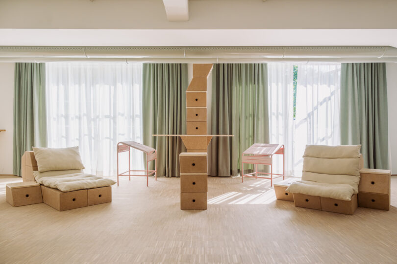 A minimalist room with light wood flooring, two cushioned chairs, a central column with stacked boxes, two desks, and large windows with sheer and green curtains