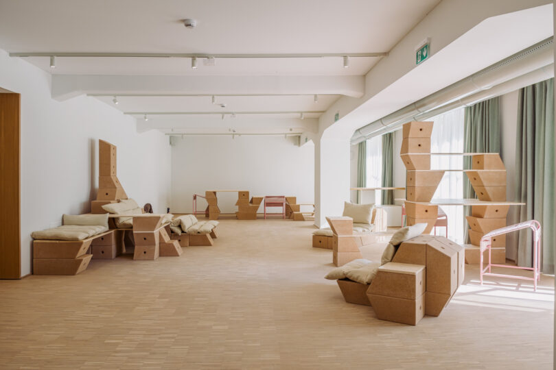 Spacious room with cork geometric furniture, neutral cushions, wooden floor, large windows with green curtains, and minimalist white walls