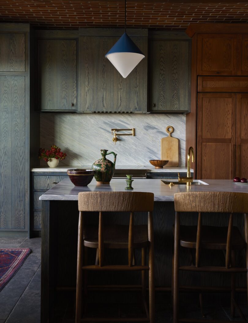 La cocina moderna cuenta con gabinetes de madera oscura, un protector contra salpicaduras de mármol, una isla con dos sillas de madera y una lámpara colgante azul y blanca en el techo.