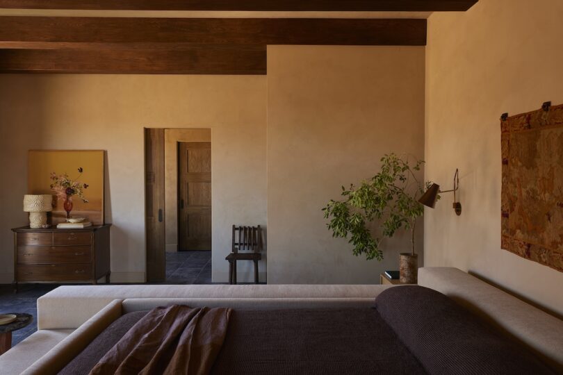 A bedroom with beige walls, wooden ceiling beams, a bed with dark bedding, a chest of drawers, a chair, a potted plant, and wall art. Warm natural light enters the room.