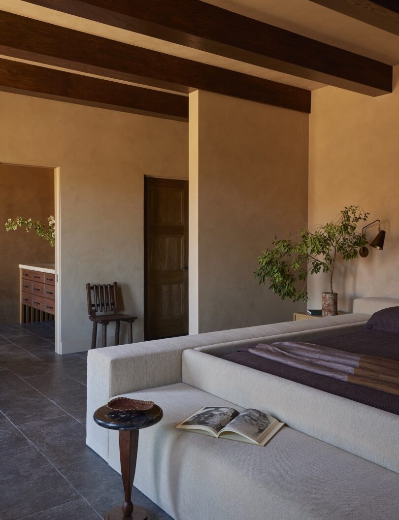 Minimalist bedroom with exposed wooden ceiling beams, neutral-toned walls, a low bed, potted plants, open book on a side table, and view into an adjacent room with a wooden bench.