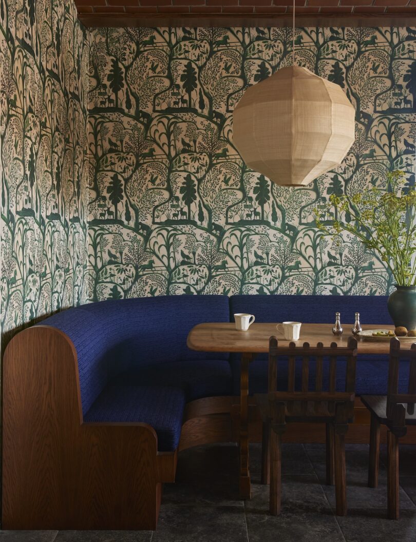 A dining nook with a curved blue upholstered bench, wooden table and chairs, green patterned wallpaper, a paper lantern light, and a vase with branches.