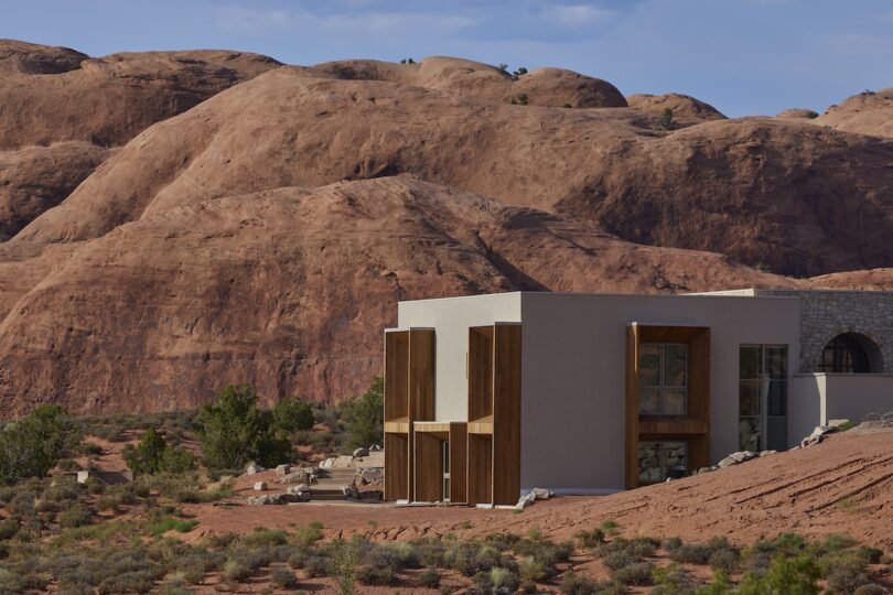 A modern building with large windows and wooden accents stands in a rocky desert landscape with reddish sandstone hills in the background.