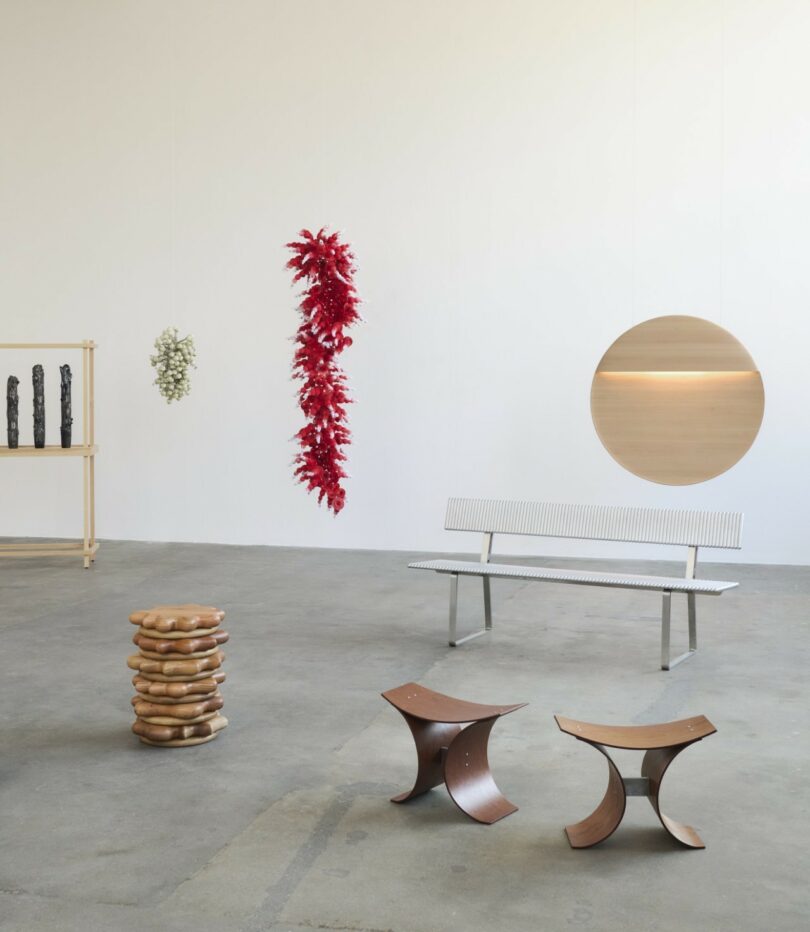 A variety of contemporary furniture and sculptural pieces are arranged in a minimalist gallery with white walls and a concrete floor