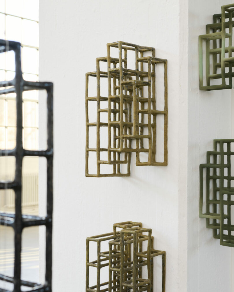 Geometric metal grid sculptures are mounted on and suspended around a white column in a sunlit gallery space with large windows