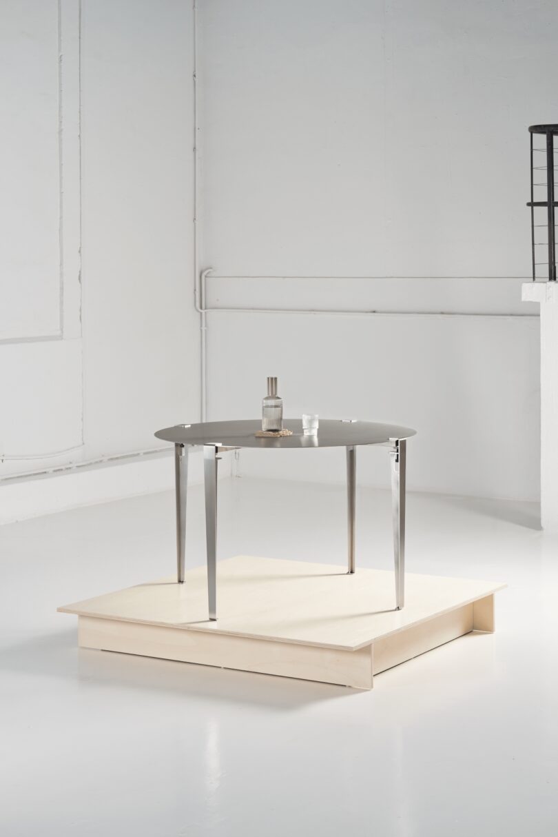 A round metal table with four legs stands on a light wooden platform. On the table are a bottle, a glass, and a small plate, as if set for someone on tiptoe in this minimal, white-painted room