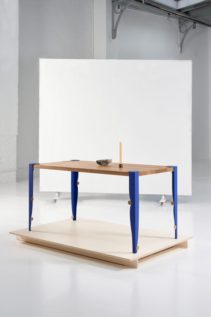 A wooden table with blue legs stands tiptoe on a light platform in a minimalistic, industrial-style room with white walls and a white backdrop