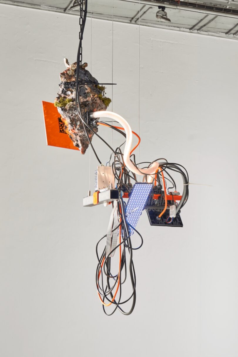 A mixed-media sculpture hangs from the ceiling, featuring tangled wires, circuit boards, LED panels, orange panels, and a rock-like object with moss, inviting viewers to tiptoe closer against a plain background