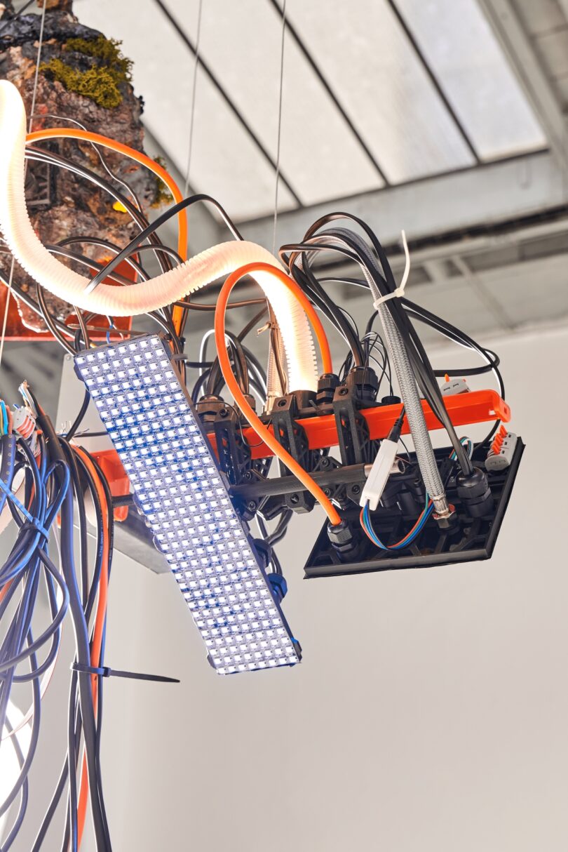 A close-up of an LED panel light and various cables and wires attached to a mechanical structure, suspended from the ceiling in an industrial setting, almost as if on tiptoe above the workspace