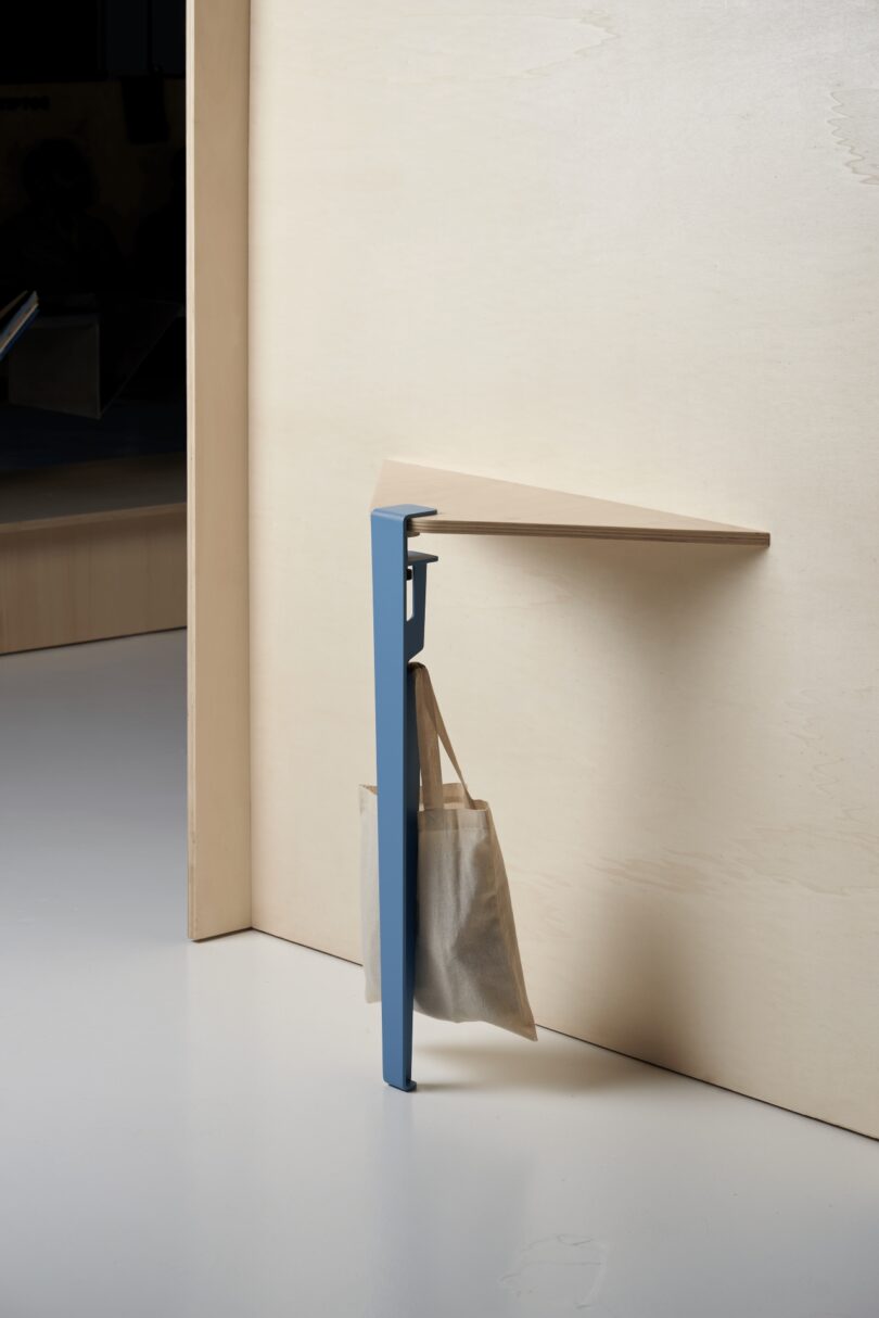 A minimalist wooden corner shelf with a blue Tiptoe metal support leg, featuring a hook holding a beige tote bag, is attached to a light wood wall