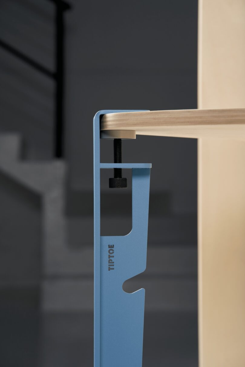 Close-up of a blue metal clamp labeled "Tiptoe" securing a wooden tabletop, with a staircase and dark background visible