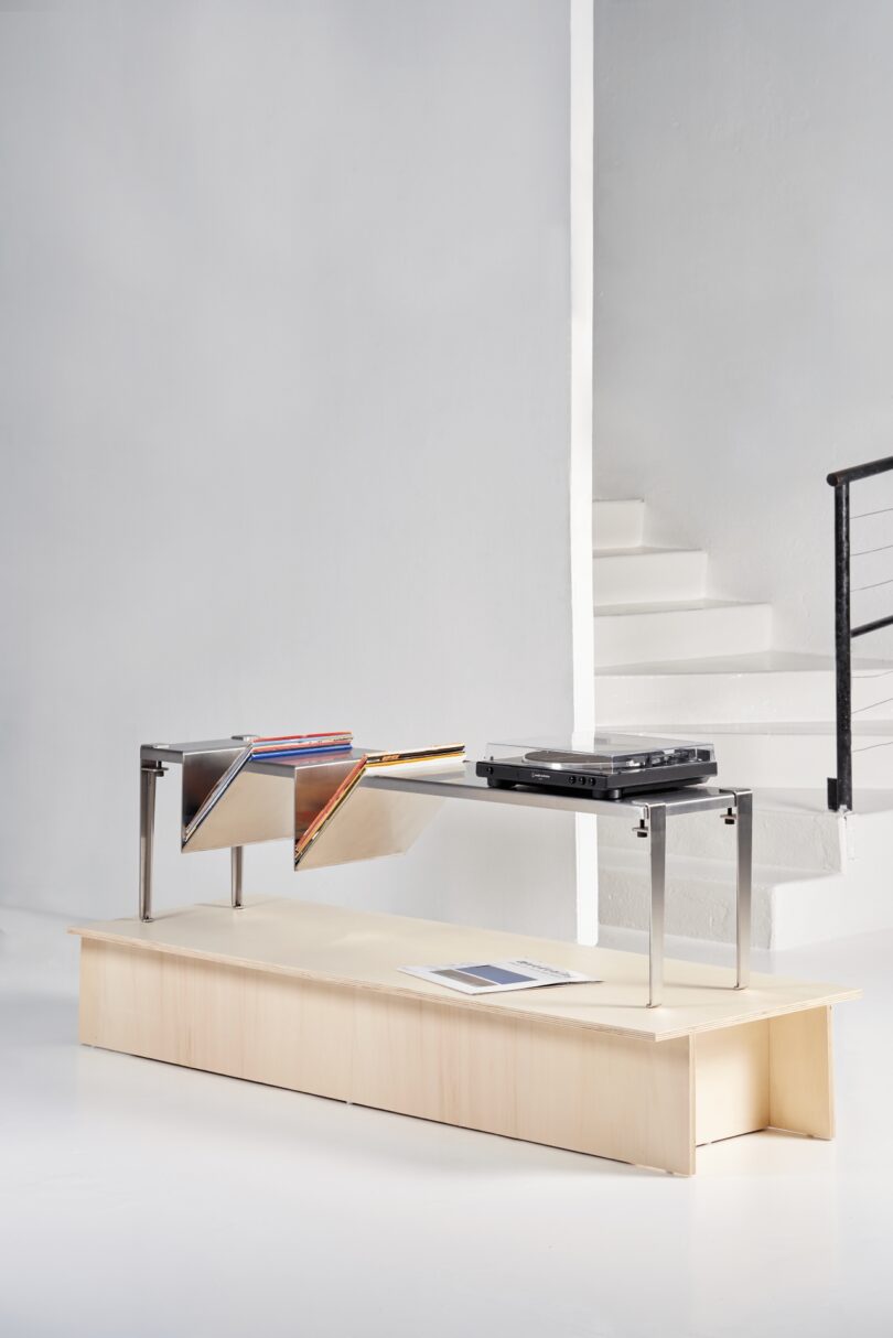 A modern wooden bench with Tiptoe legs and a metal record storage rack holding vinyl records and a turntable, set against a white staircase and minimalist interior