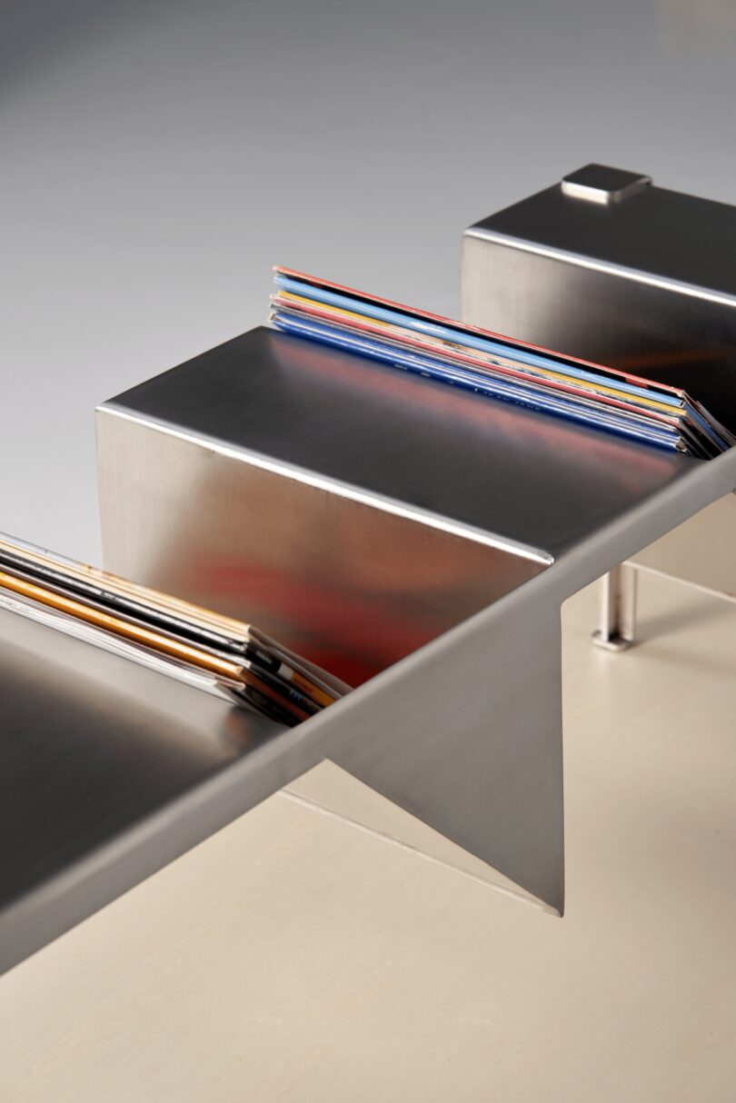 A modern metal shelf stands on tiptoe, holding several stacks of colorful vinyl records, positioned at an angle on a light-colored floor