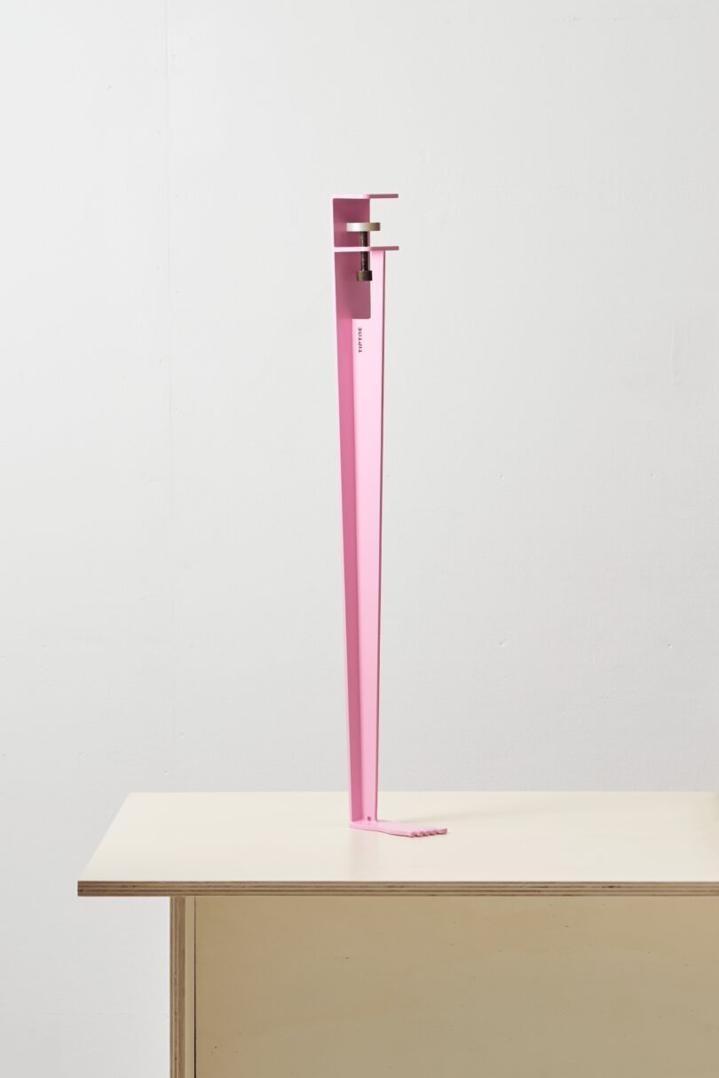 A tall, narrow, pink metal object stands on tiptoe with a small clamp at the top, poised upright on a light-colored table against a plain white wall
