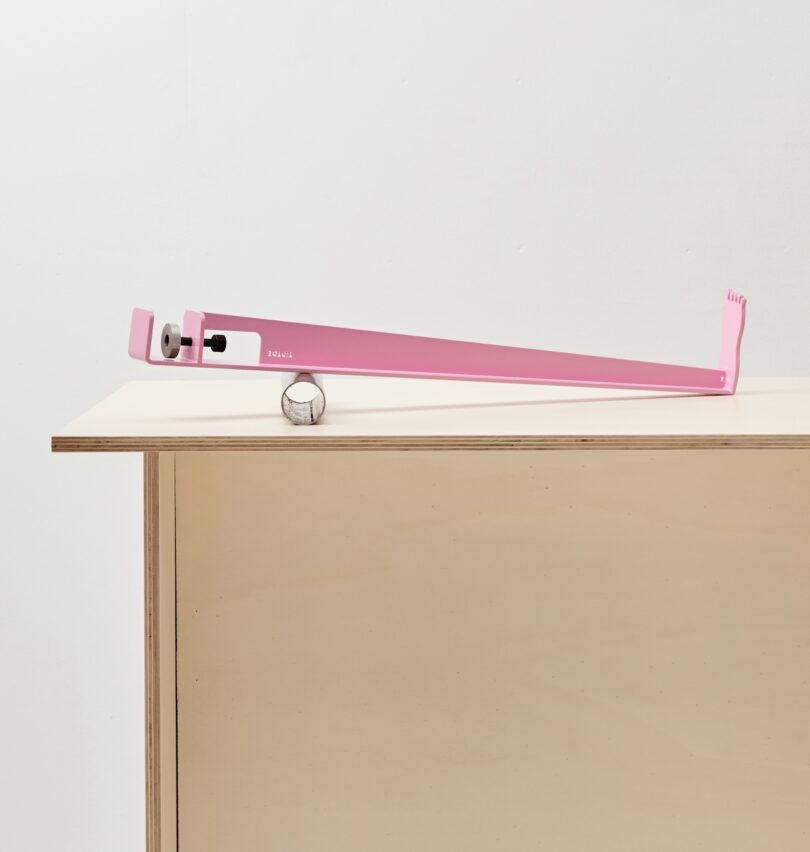 A long pink metal tool with tiptoes across a light wood table, with a small silver egg-shaped object resting nearby