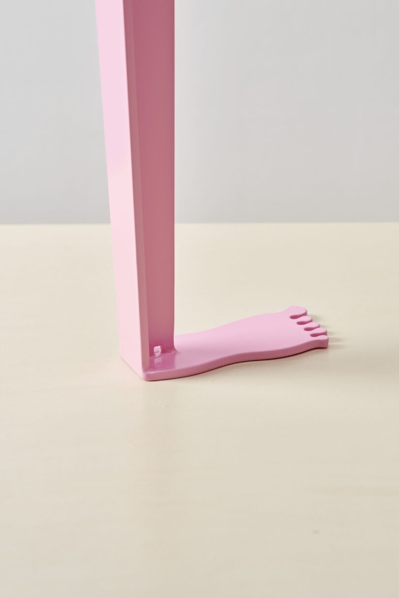 A pink rectangular table leg with a flat base shaped like a human foot stands on tiptoe atop a light-colored surface
