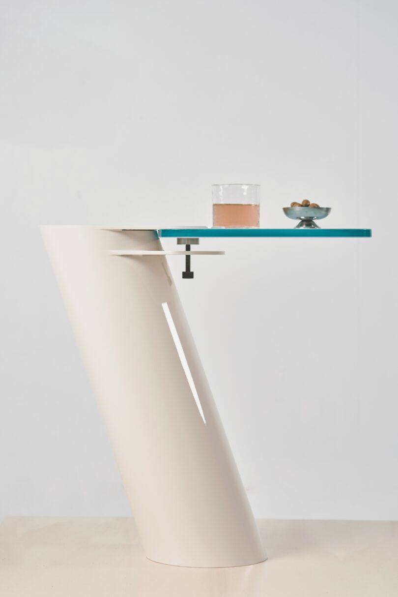 A modern, angled white Tiptoe table with a glass top holds a drink in a clear glass and a small bowl of olives against a plain background
