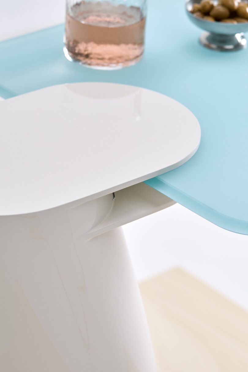 Close-up of a modern light blue table just within tiptoe reach, featuring a white chair, a glass of pink liquid, and a small bowl of green olives