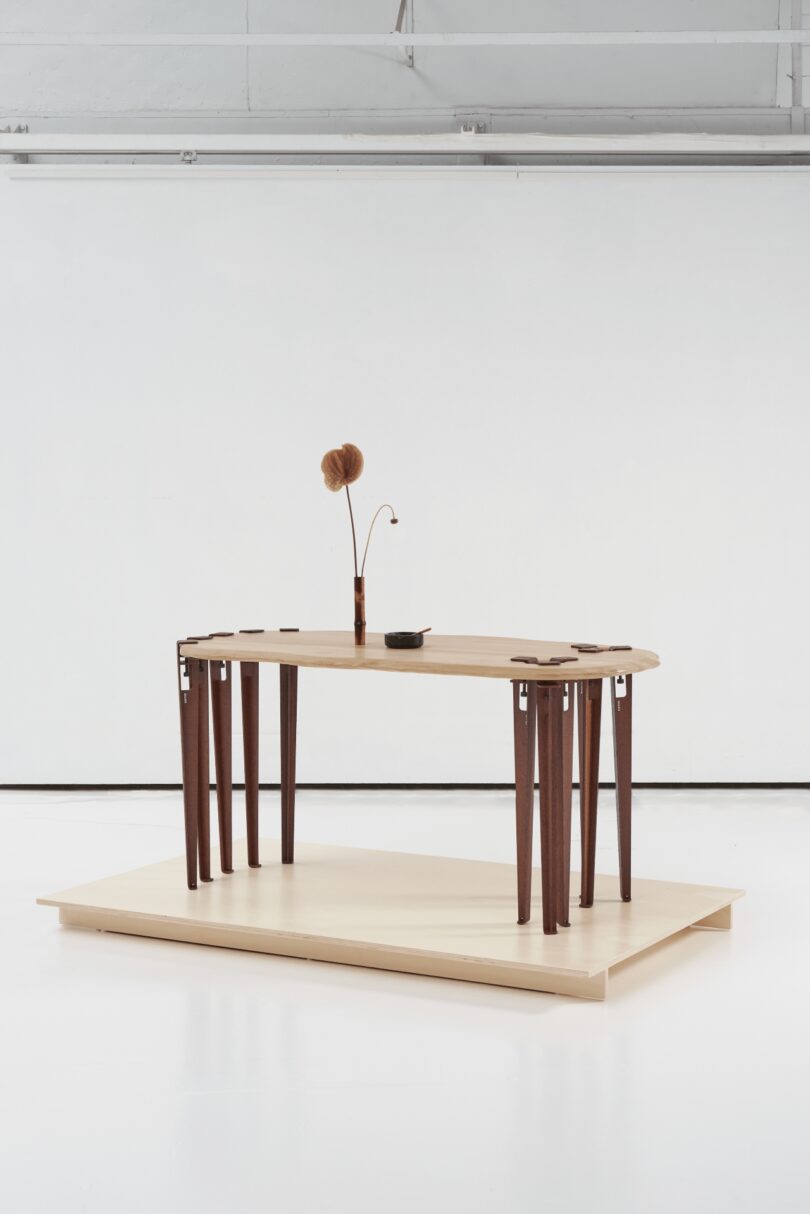 A modern wooden table with multiple dark tiptoe-style legs, decorated with minimalist vases and dried flowers, set on a light platform in a white, industrial-style room